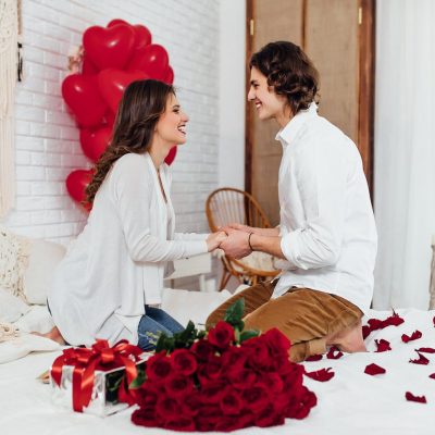 happy couple holding hands, bouquet of red roses and gift box on the bed